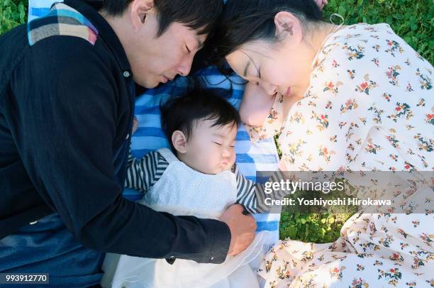father and mother sleeping with toddler girl ( 6-11 months ) on lawn at park under sunshine - stadtteil koto stock-fotos und bilder