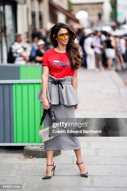 Guest wears yellow glasses, a red FILA t-shirt with a printed logo, a gray skirt, gray heels shoes, a silver shiny Diorama Dior bag , outside...