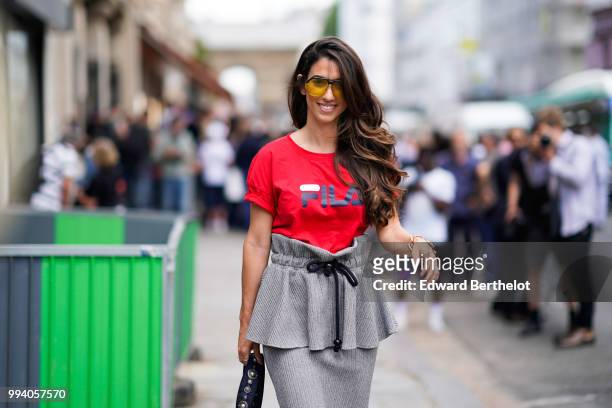 Guest wears yellow glasses, a red FILA t-shirt with a printed logo, a gray skirt, gray heels shoes, a silver shiny Diorama Dior bag , outside...