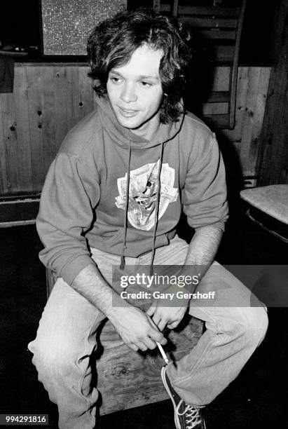 View of American Rock, County, and Folk musician John Cougar, a cigarette in his handm as he sits on a wooden box backstage at the nightclub, My...