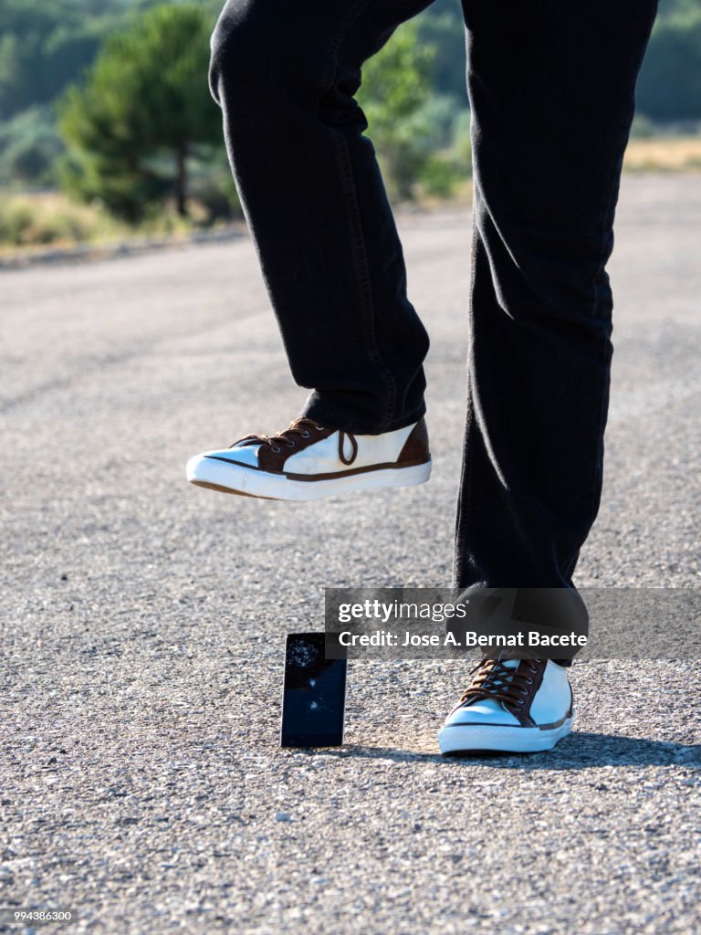 Slipper of white color of a person squishing a mobile phone against the soil, outdoors.