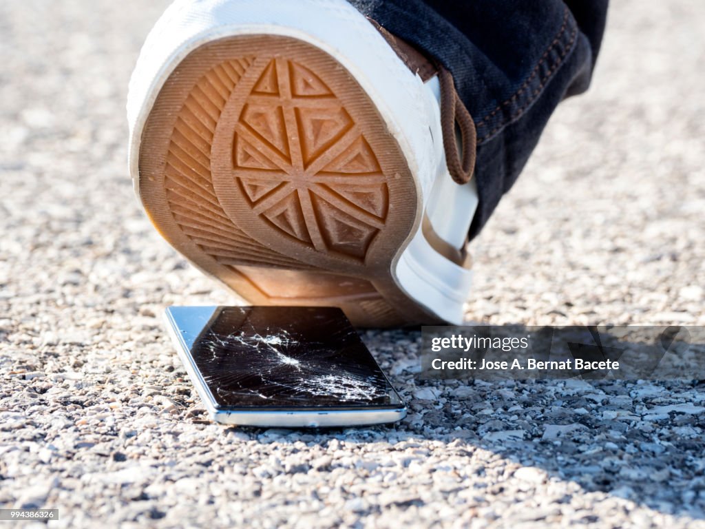 Slipper of white color of a person squishing a mobile phone against the soil, outdoors.