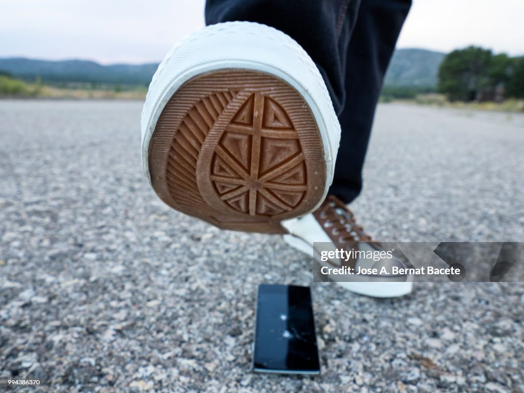 Slipper of white color of a person squishing a mobile phone against the soil, outdoors.