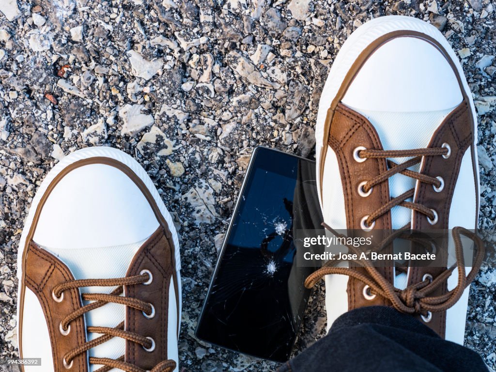 Slipper of white color of a person squishing a mobile phone against the soil, outdoors.