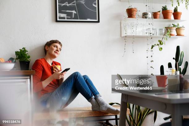 woman sitting in kitchen, drinking coffee and checking smartphone messages - woman drinking phone kitchen stockfoto's en -beelden