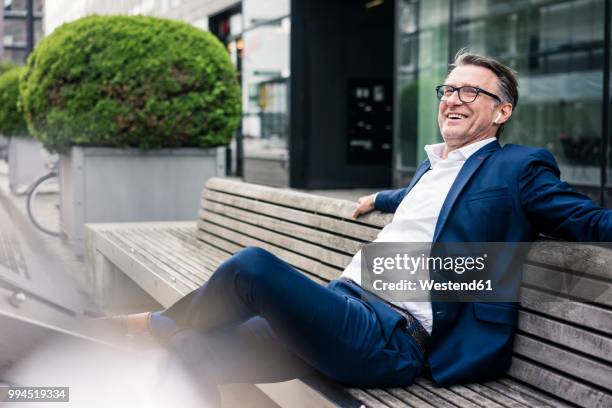 happy mature businessman sitting on bench outdoors wearing earbuds - bank exterior stock-fotos und bilder
