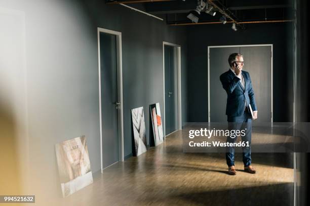 mature businessman standing on office floor with paintings using cell phone - kunsthändler stock-fotos und bilder