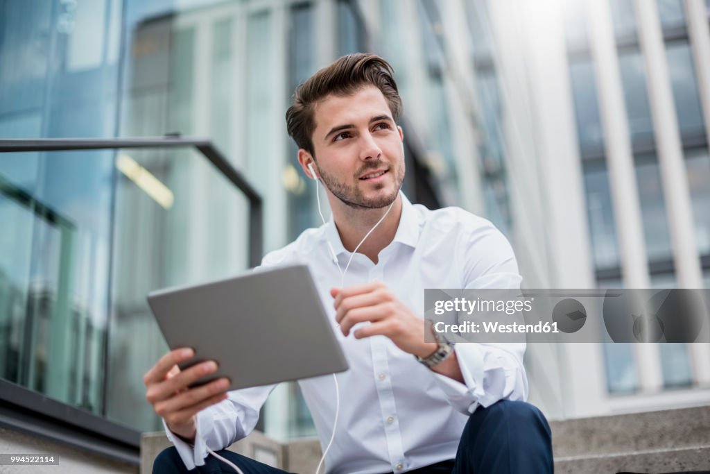 Smiling businessman sitting on stairs with earbuds and tablet