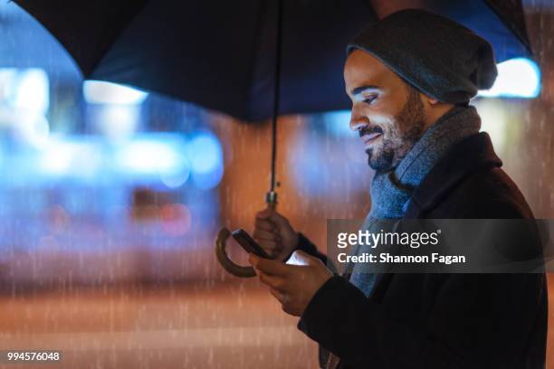 street portrait of a young man holding mobile phone on a rainy day - man with umbrella stockfoto's en -beelden