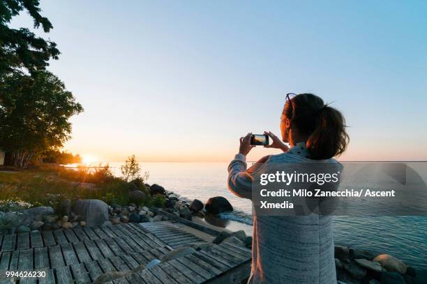 woman pauses on lakeshore at sunrise, takes pic - technophiler mensch stock-fotos und bilder