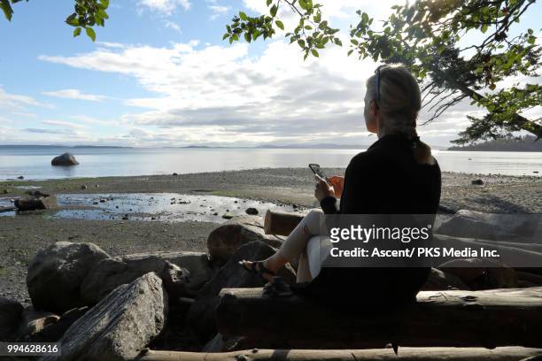 woman relaxes on shoreline log, look out at calm sea - technophile stock pictures, royalty-free photos & images