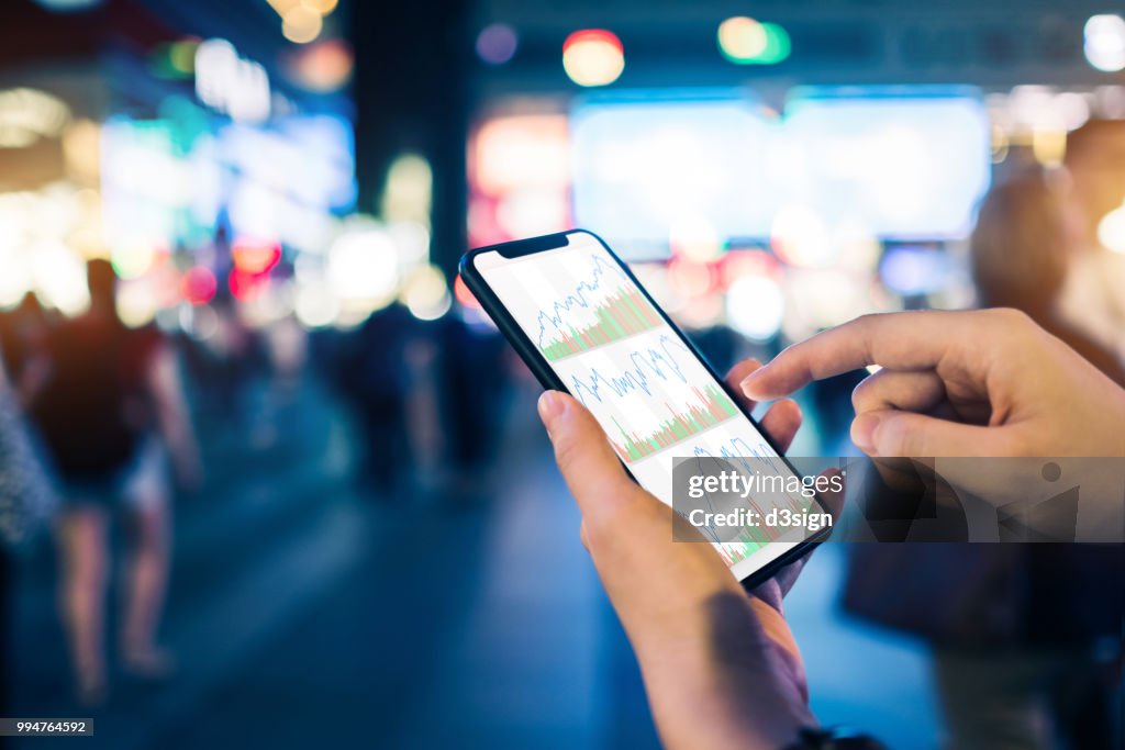 Close up of human hands checking financial stock charts on smartphone in busy city street, against neon commercial sign at night