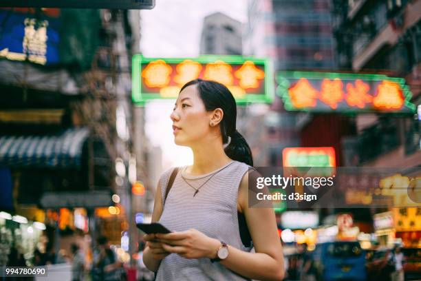 beautiful young female traveller searching for direction on smartphone on busy city street, against colourful neon commercial sign and city buildings - temple street market stock pictures, royalty-free photos & images