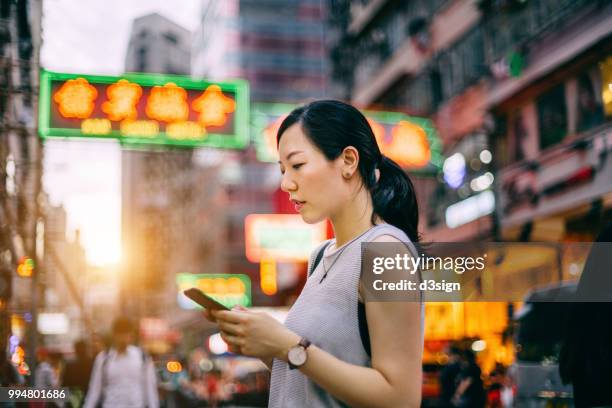 beautiful young woman using smartphone in busy city street, against colourful neon commercial sign and city buildings - temple street market stock pictures, royalty-free photos & images