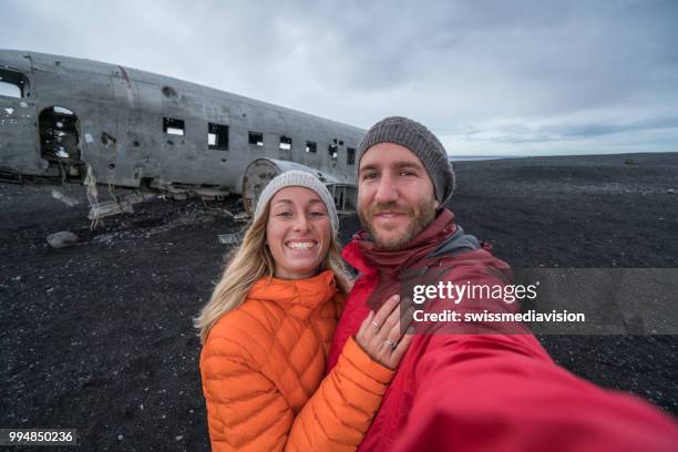 junges paar stehend durch flugzeug wrack am schwarzen sandstrand unter selfie porträt sehenswürdigkeit zu besuchen in island und ist mit dem wrack - swissmediavision stock-fotos und bilder