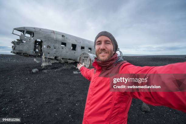 jeune homme debout par avion épave sur la plage de sable noir, tenant un lieu célèbre portrait de selfie à visiter en islande et posent avec l’épave - swissmediavision photos et images de collection