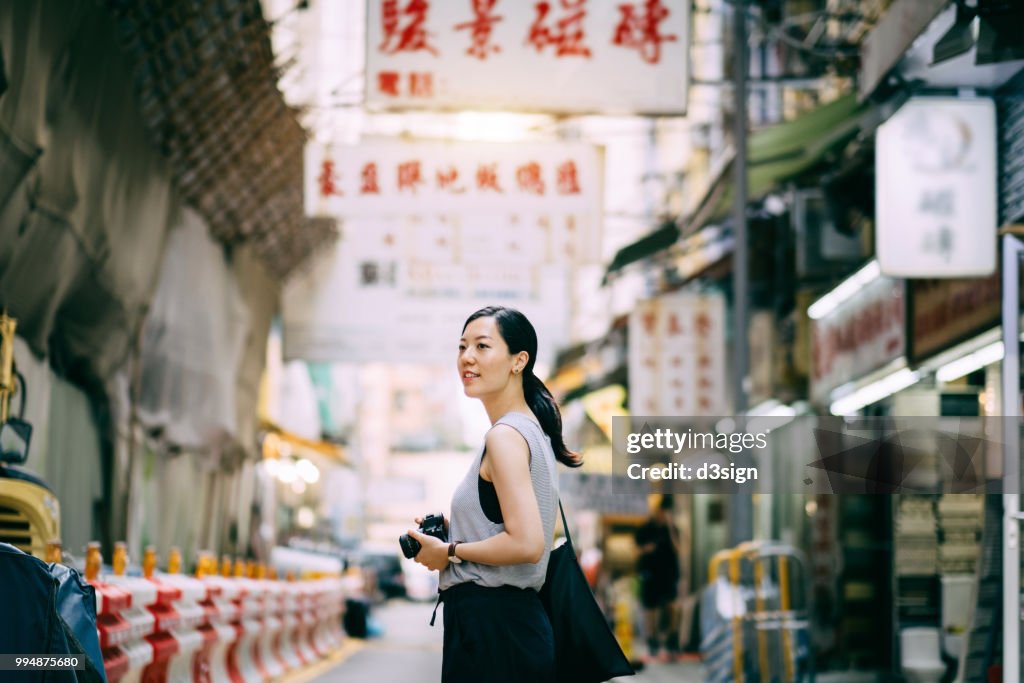 Beautiful young woman carrying camera exploring and walking through local city street