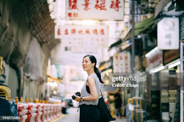 beautiful young woman carrying camera exploring and walking through local city street - chinese ethnicity stock pictures, royalty-free photos & images