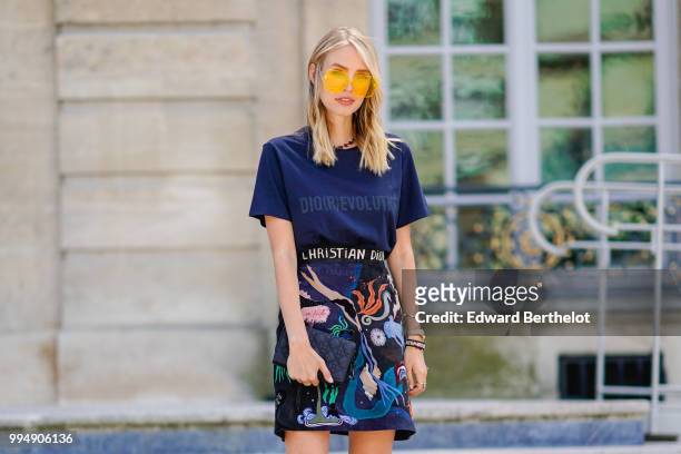 Leonie Hanne wears a dark blue Dior Evolution t-shirt, a black clutch, a flower print skirt, yellow glasses, outside Dior, during Paris Fashion Week...