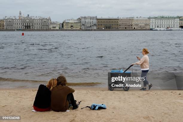 Couple sits as a woman walks with a baby carriage along the Neva River in Saint Petersburg, Russia on July 09, 2018.