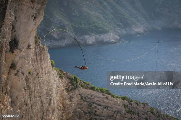 Members of the rope jump french team Pyrenaline jump from atop the rugged rocks overlooking the azure waters, Ionian Islands, Zakynthos, Greece.