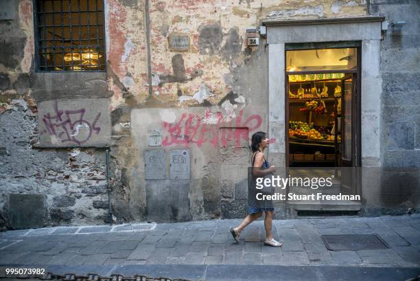 Woman walks past a grocers shop in the Old centre of Genoa, Italy. The heart of medieval Genoa is bounded by ancient city gates Porta dei Vacca and...