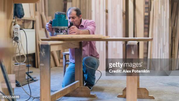 el hombre mayor de 65 años de edad trabajando con la fresadora de mano eléctrica. carpinterías de madera en la fábrica de muebles pequeños. - alex potemkin or krakozawr fotografías e imágenes de stock