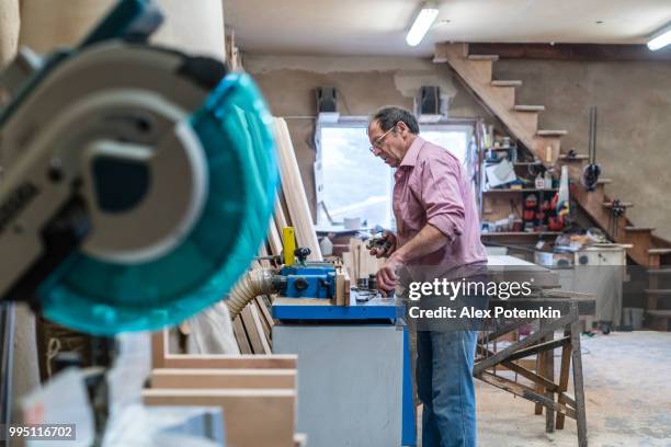 el hombre mayor de 60 años de edad, carpintero y sashman, artesano, trabajando con la fresadora industrial en la pequeña fábrica de madera - alex potemkin or krakozawr fotografías e imágenes de stock