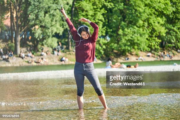 happy young woman listening music with headphones and cell phone dancing at riverside - río isar fotografías e imágenes de stock