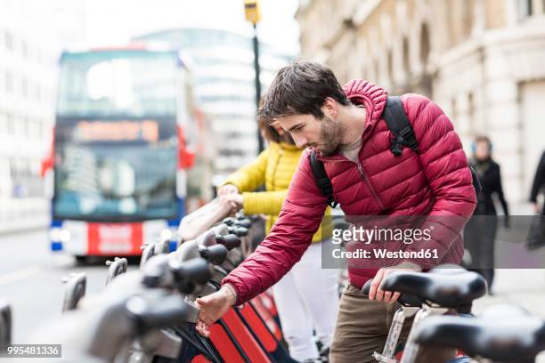 uk, london, young man renting bicycle from bike share stand in city - london bikes stock pictures, royalty-free photos & images