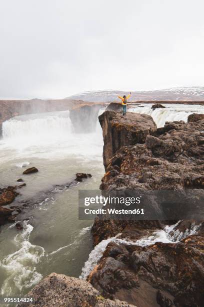 iceland, woman standing at godafoss waterfall with raised arms - northeast iceland stockfoto's en -beelden