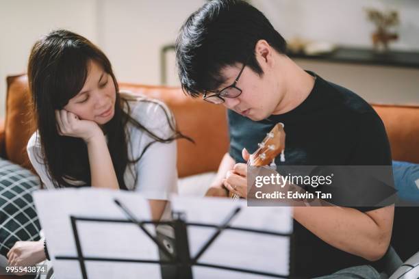 un maschio cinese asiatico insegnamento di una femmina cinese asiatica su ukulele in soggiorno - plucking an instrument foto e immagini stock