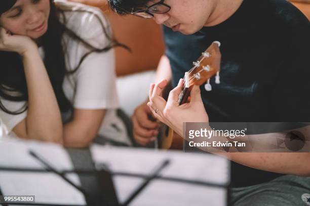un maschio cinese asiatico insegnamento di una femmina cinese asiatica su ukulele in soggiorno - plucking an instrument foto e immagini stock