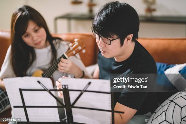 un maschio cinese asiatico insegnamento di una femmina cinese asiatica su ukulele in soggiorno - plucking an instrument foto e immagini stock