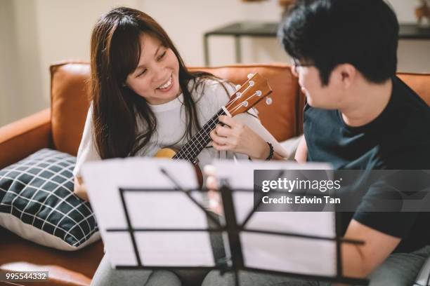 un maschio cinese asiatico insegnamento di una femmina cinese asiatica su ukulele in soggiorno - plucking an instrument foto e immagini stock