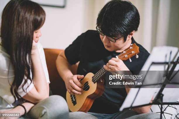 un maschio cinese asiatico insegnamento di una femmina cinese asiatica su ukulele in soggiorno - plucking an instrument foto e immagini stock