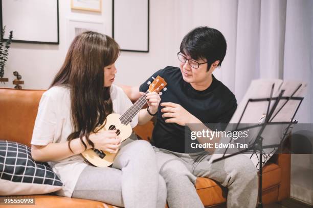 un maschio cinese asiatico insegnamento di una femmina cinese asiatica su ukulele in soggiorno - plucking an instrument foto e immagini stock