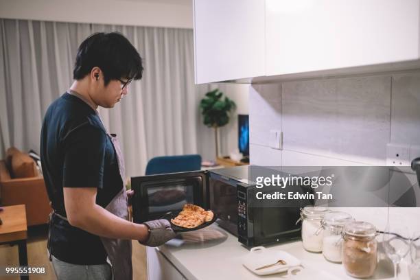 einen asiatischen chinesischen mann nehmen eine pizza aus der mikrowelle in der küche mit handschuh - mikrowelle stock-fotos und bilder