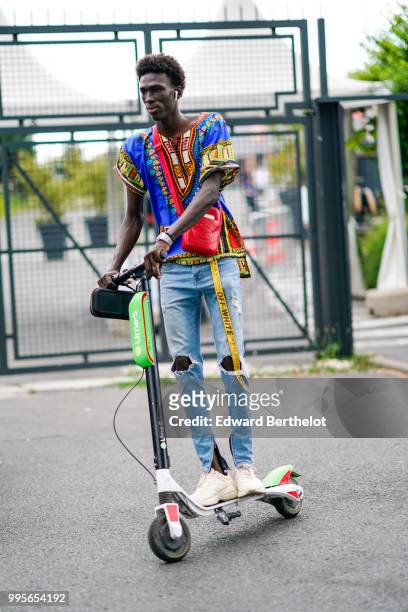 Model wears a multicolor printed top, a yellow off-white belt, blue ripped jeans, white sneakers shoes, a red leather shoulder strap bag, and rides...