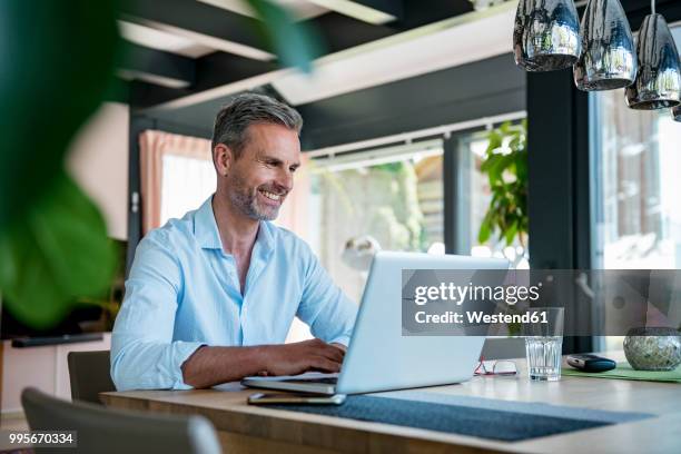 smiling mature man at home using a laptop at table - 40s laptop stockfoto's en -beelden