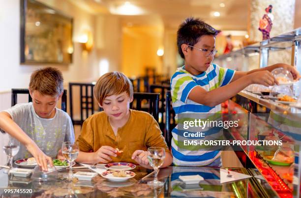 3 boys of diverse ethnicities enjoying all you can eat asian food in running sushi restaurant - sushi train stock pictures, royalty-free photos & images