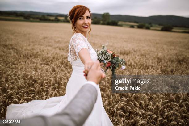 bella giovane sposa che si tiene per mano del marito sul campo di grano il giorno del suo matrimonio - prospettiva del fotografo foto e immagini stock