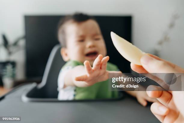 crying baby sitting on highchair reaching for a slice of fruit from mother's hand - toddler crying stock pictures, royalty-free photos & images