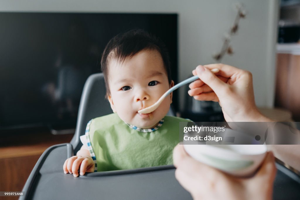 Mother's hand feeding baby girl food on high chair at home