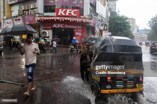 Pedestrian holding an umbrella crosses a road in the rain as an auto-rickshaw drives through a waterlogged street in front of a Yum! Brands Inc. KFC...