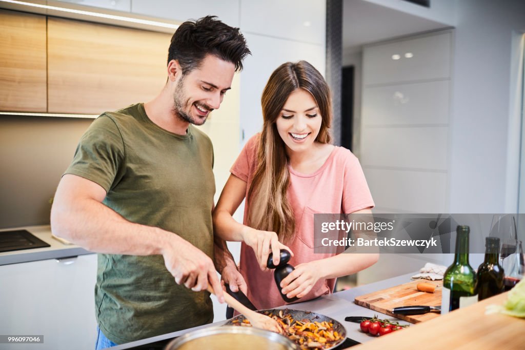 Linda pareja feliz cocinando juntos y añadir especias a la comida, reír y pasar tiempo juntos en la cocina