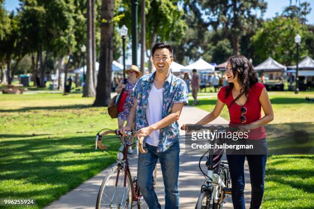 los angeles couple wheeling bikes through park durning farmer's market - pasadena california stock pictures, royalty-free photos & images