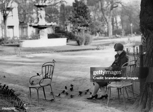 Woman sitting on a chair in a park , next to her: sparrows on the ground and on a chair. Photograph. Paris. Around 1930.