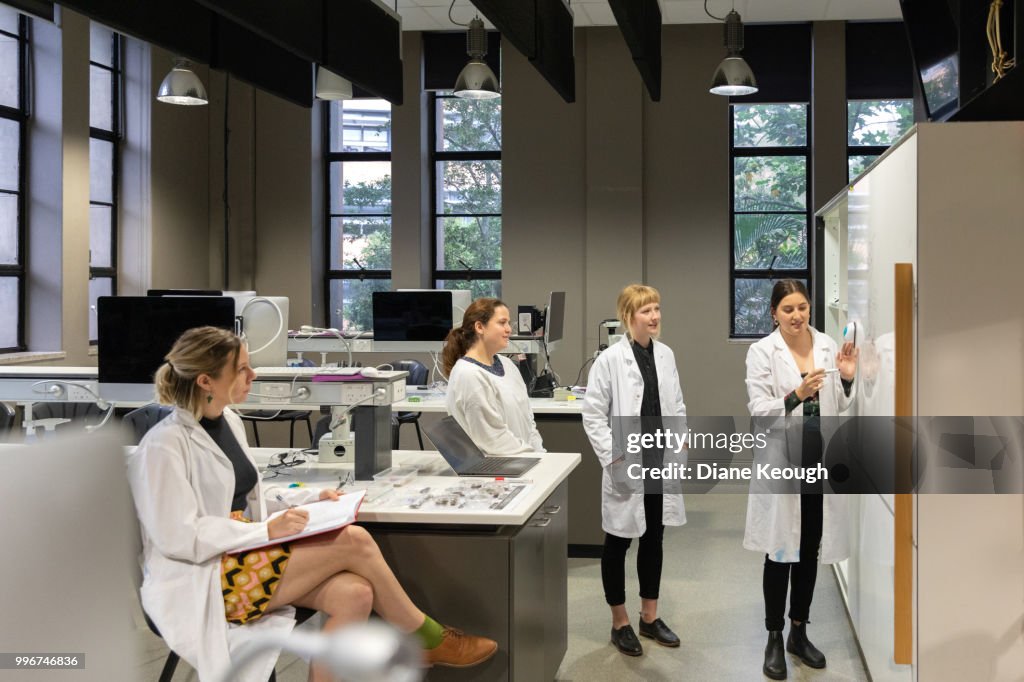 4 female scientists , 3 standing and 1 sitting show full length in a laboratory with one teaching the research from a whiteboard. They are all looking toward the whiteboard and wearing lab coats. Wider view with the laboratory area shown behind them.