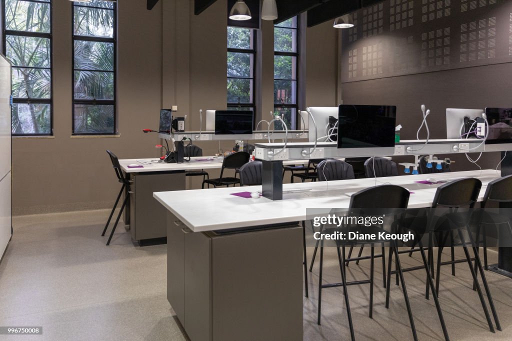 Side view of a clean teaching laboratory. Contains 2 long desks running right to left with high chairs and computer equipment and windows in the background.